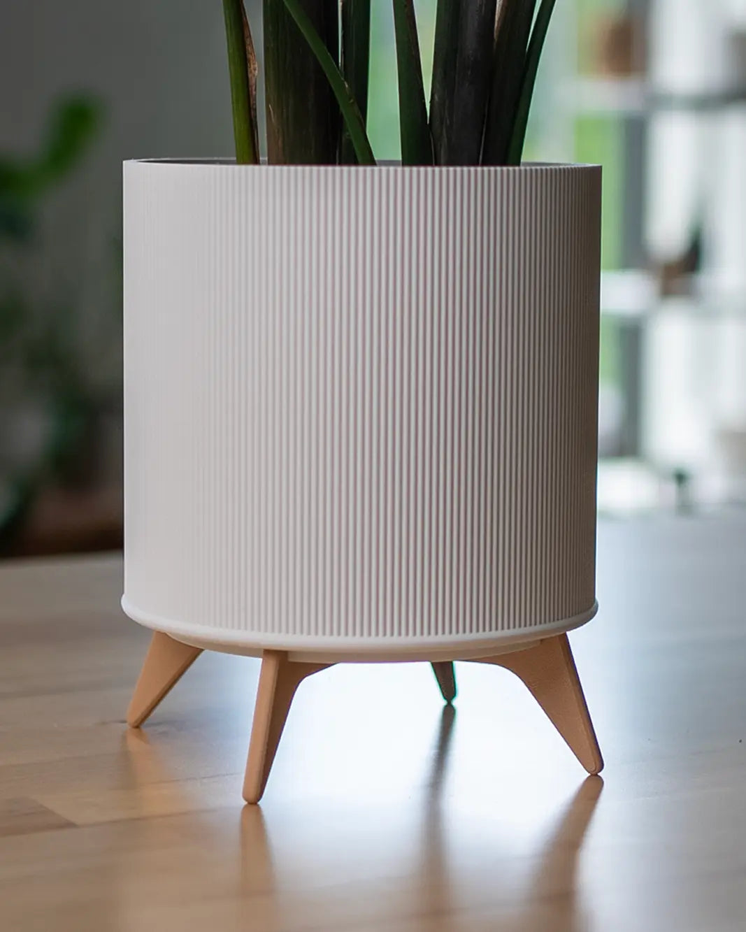 Der Evoliv „The Tyler“ – Moderner Pflanztopf mit Ständer in Weiß mit vertikalen Rillen steht stilvoll auf einem Holztisch. Grüne Zimmerpflanzenblätter ragen empor, während der Hintergrund sanft Pflanzen und Regale andeutet.