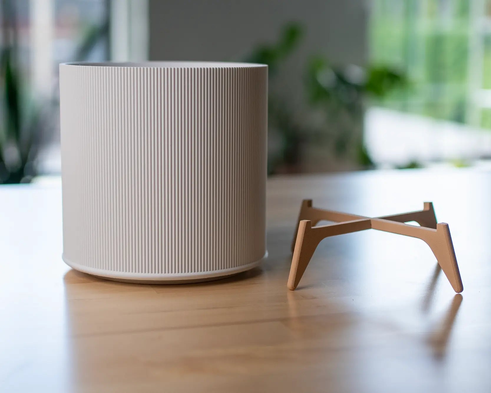 Der Evoliv „The Tyler“ – moderner Pflanztopf mit Ständer – in Weiß mit vertikalen Rillen steht auf einer Holzoberfläche neben einem kleinen, hellbraunen Pflanzenständer. Im Hintergrund sind unscharfe Zimmerpflanzen und ein Fenster sichtbar.