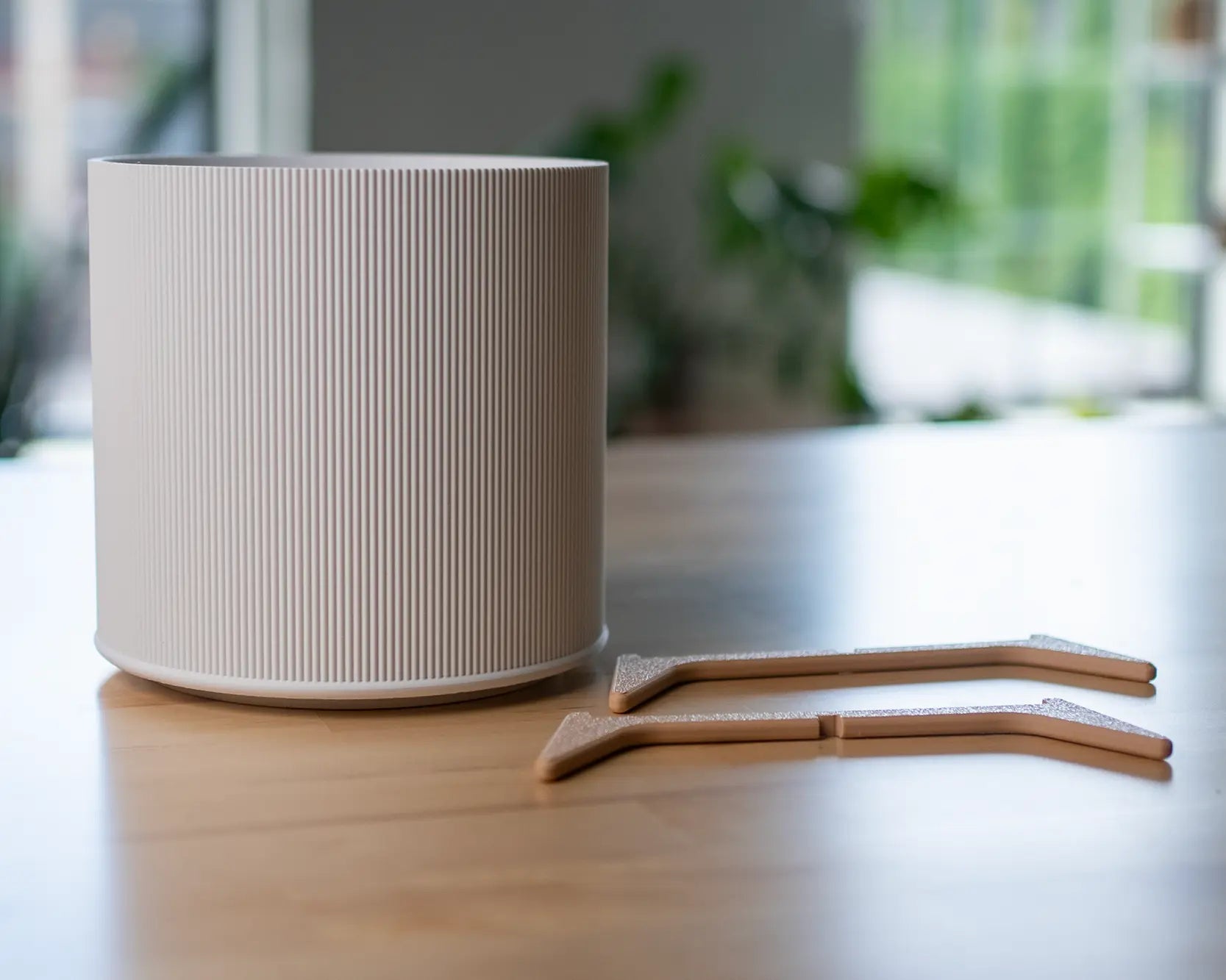 Der weiße Evoliv „The Tyler“ – Moderner Pflanztopf mit Ständer steht auf einem Holztisch neben zwei beigen Pflanzensteckern, im Hintergrund Zimmerpflanzen und Fenster unscharf zu sehen.
